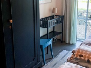 Um quarto com uma porta escura, uma cadeira azul, uma cama com almofada estampada e uma janela com vista para rochas e vegetação.