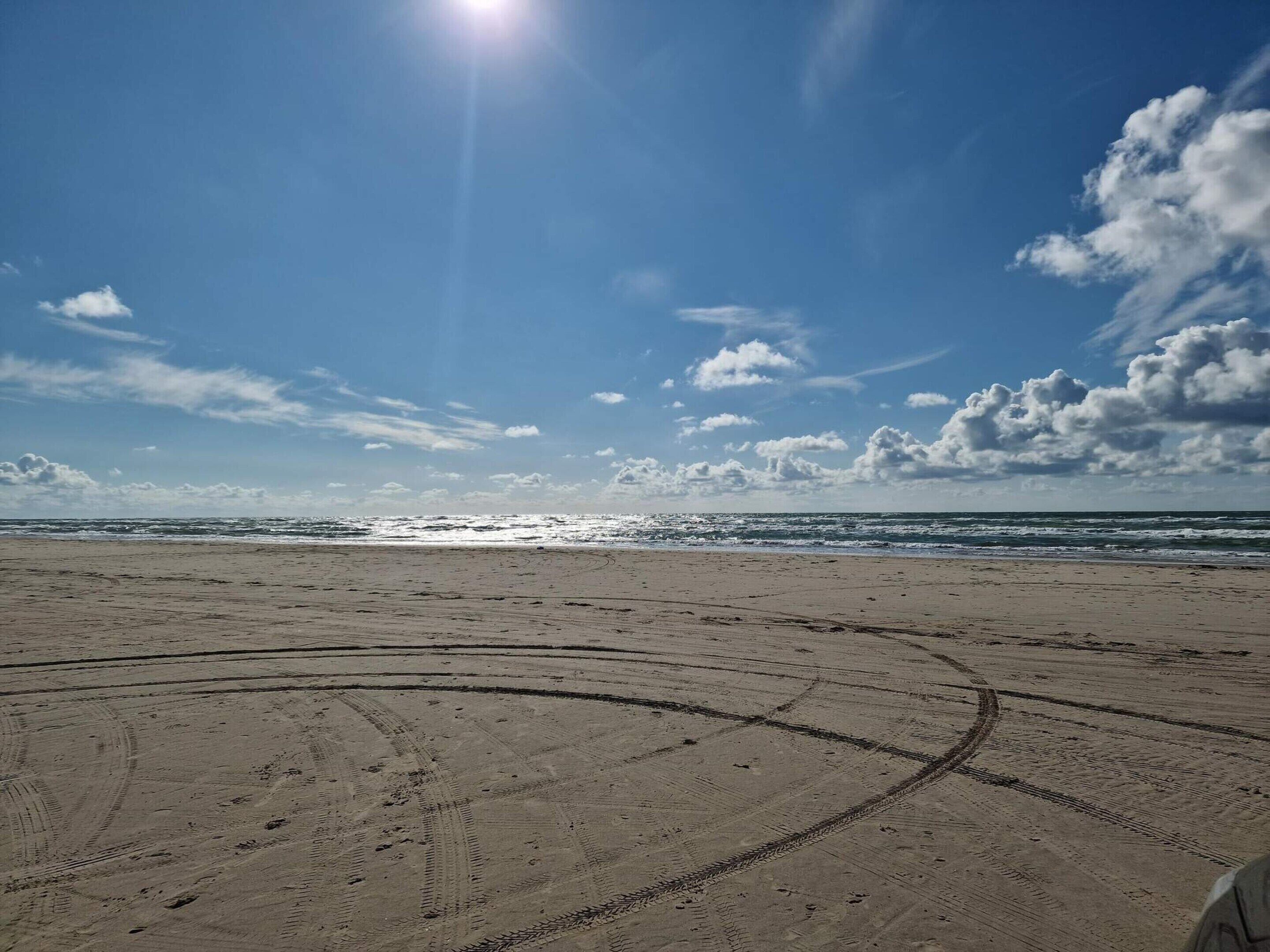 A beach with tire tracks leading to the water, a clear sky, and the sun shining.