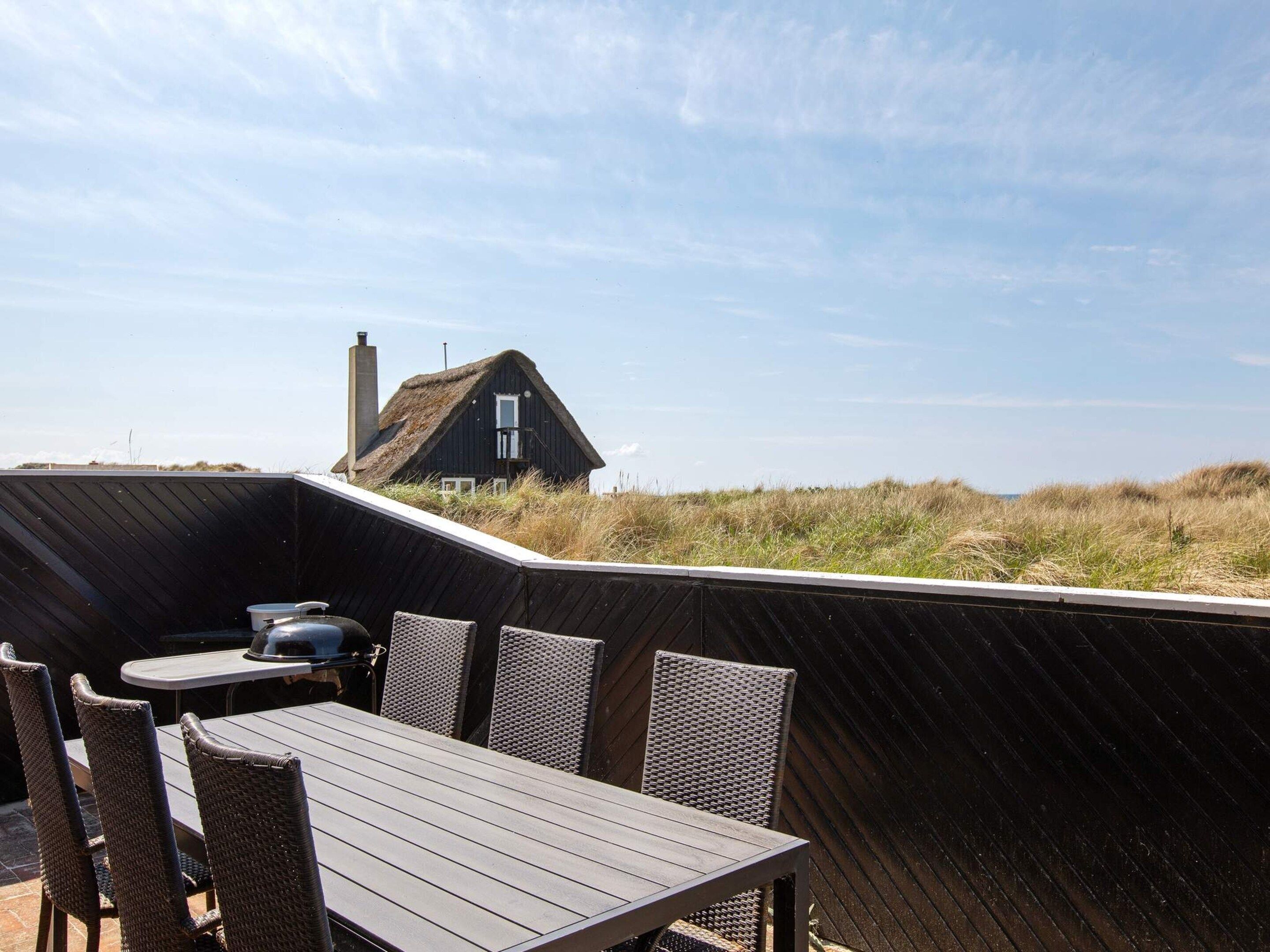 Une terrasse en bois avec une table et des chaises, d’où l’on a vue sur une maison et des dunes.