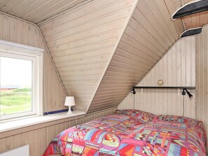 A bedroom with a sloped ceiling, a bed with a colorful quilt, a bedside lamp, and a window with a view of greenery.