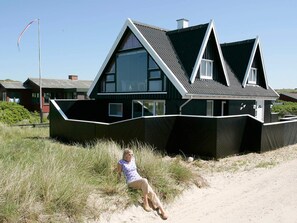 A modern house with a unique angular design, large windows, and a sandy surrounding.