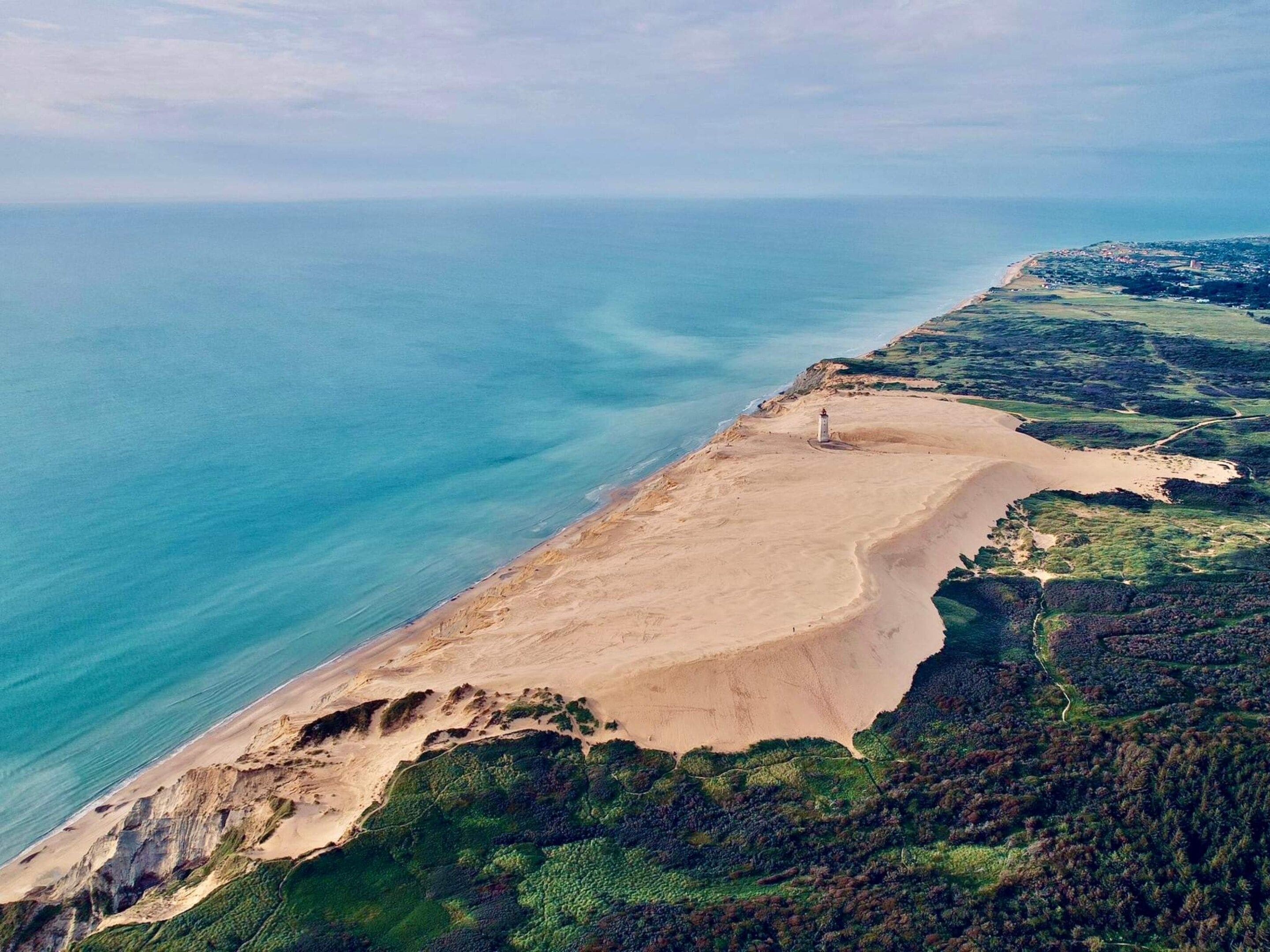 Vista aérea de un paisaje costero con una playa arenosa, un bosque verde denso y un faro.