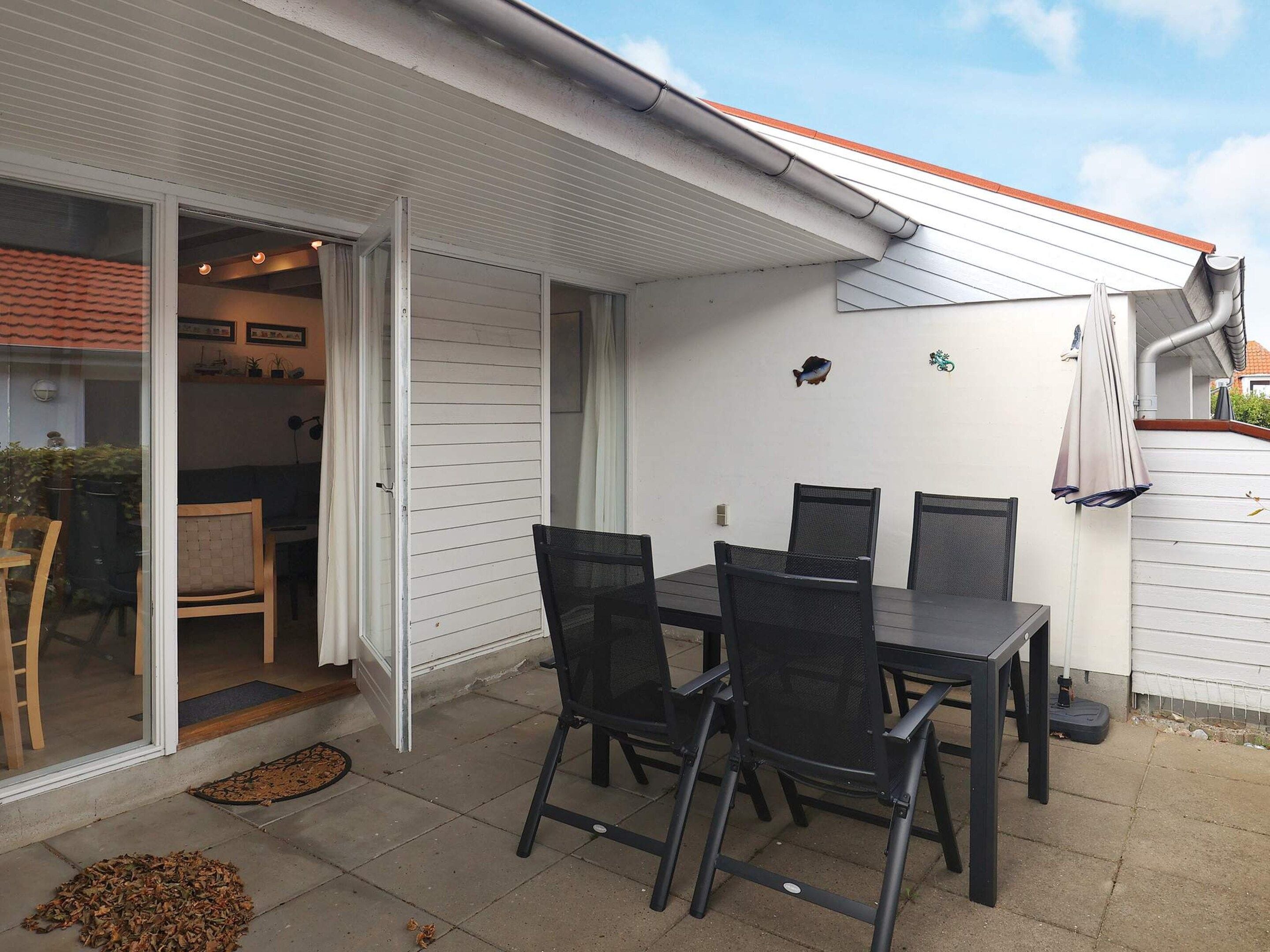 Une terrasse extérieure avec une table et des chaises, une porte-fenêtre coulissante donnant sur un espace intérieur, et un petit élément décoratif représentant un poisson sur le mur.