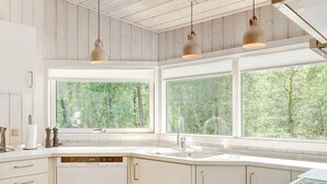 A modern kitchen with white cabinetry, a built-in dishwasher, and large windows offering a view of the forest.