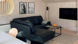 A modern living room with a black sectional sofa, a dining table with chairs, a flat-screen TV mounted on the wall, and a decorative pendant light.
