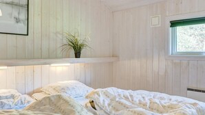 A bedroom with a wooden wall, a window, a bed with floral bedding, a shelf with a plant, and a framed picture.
