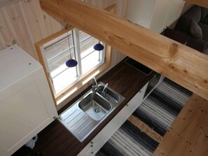 Eine Küche mit Spüle aus Edelstahl, Holzbalken, einem Fenster mit Jalousien und einem Streifen Teppich.