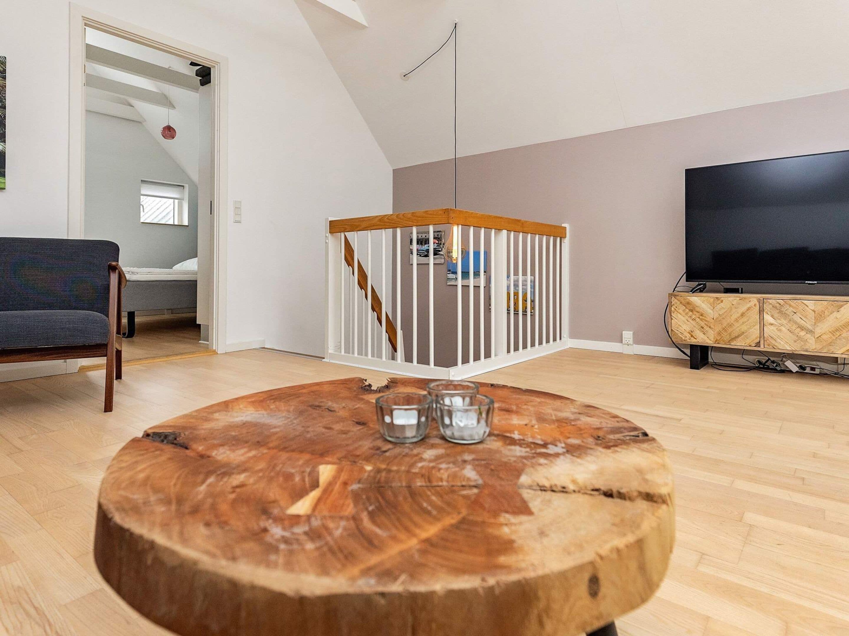 Ein modernes Wohnzimmer mit einem runden Holztisch, einem Fernseher auf einem Holzschrank und einer Treppe mit weißer Geländer.