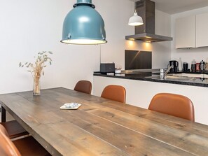A modern kitchen with a wooden dining table, two pendant lights, and a vase with dried plants.
