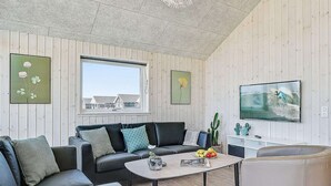 A modern living room with a sofa, a coffee table, a TV, and a window with a view of houses. - Holiday Home in Kappeln (Kappeln)