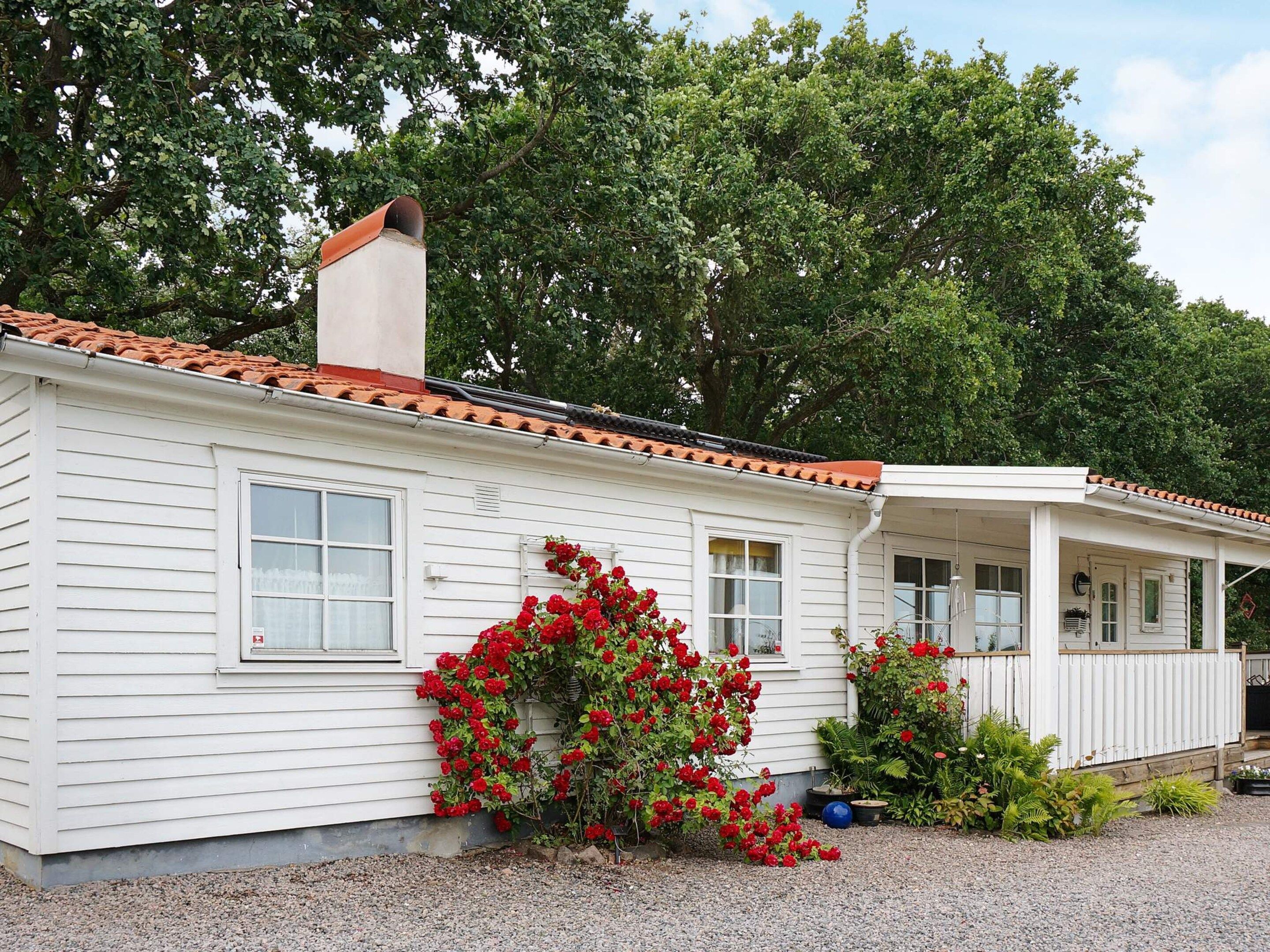 A single-story house with a white exterior, a red-tiled roof, and a chimney. There is a small porch with a white railing, and a garden with red flowers in front.