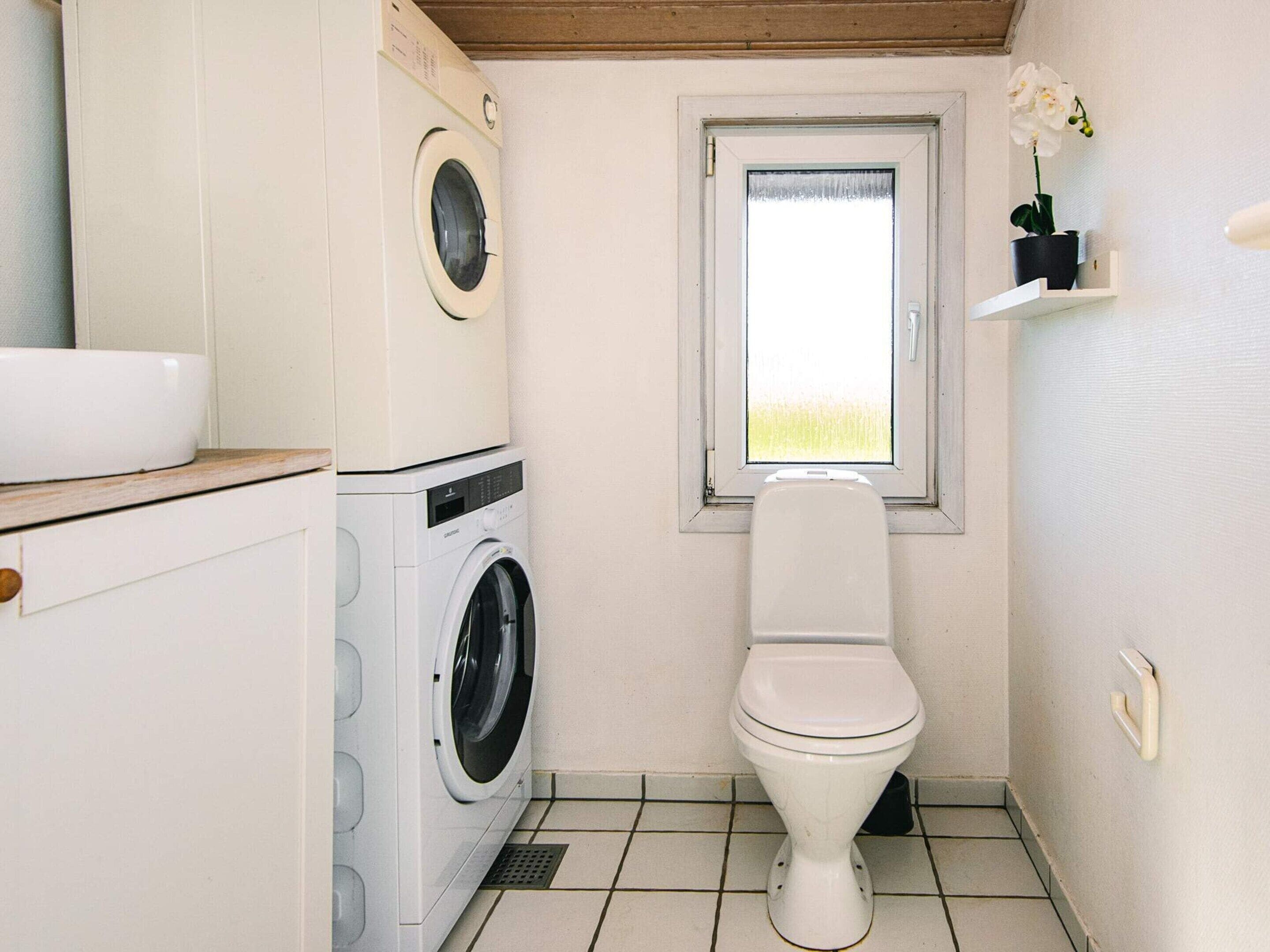 A compact bathroom with a washing machine, toilet, and a window with a view of a field.