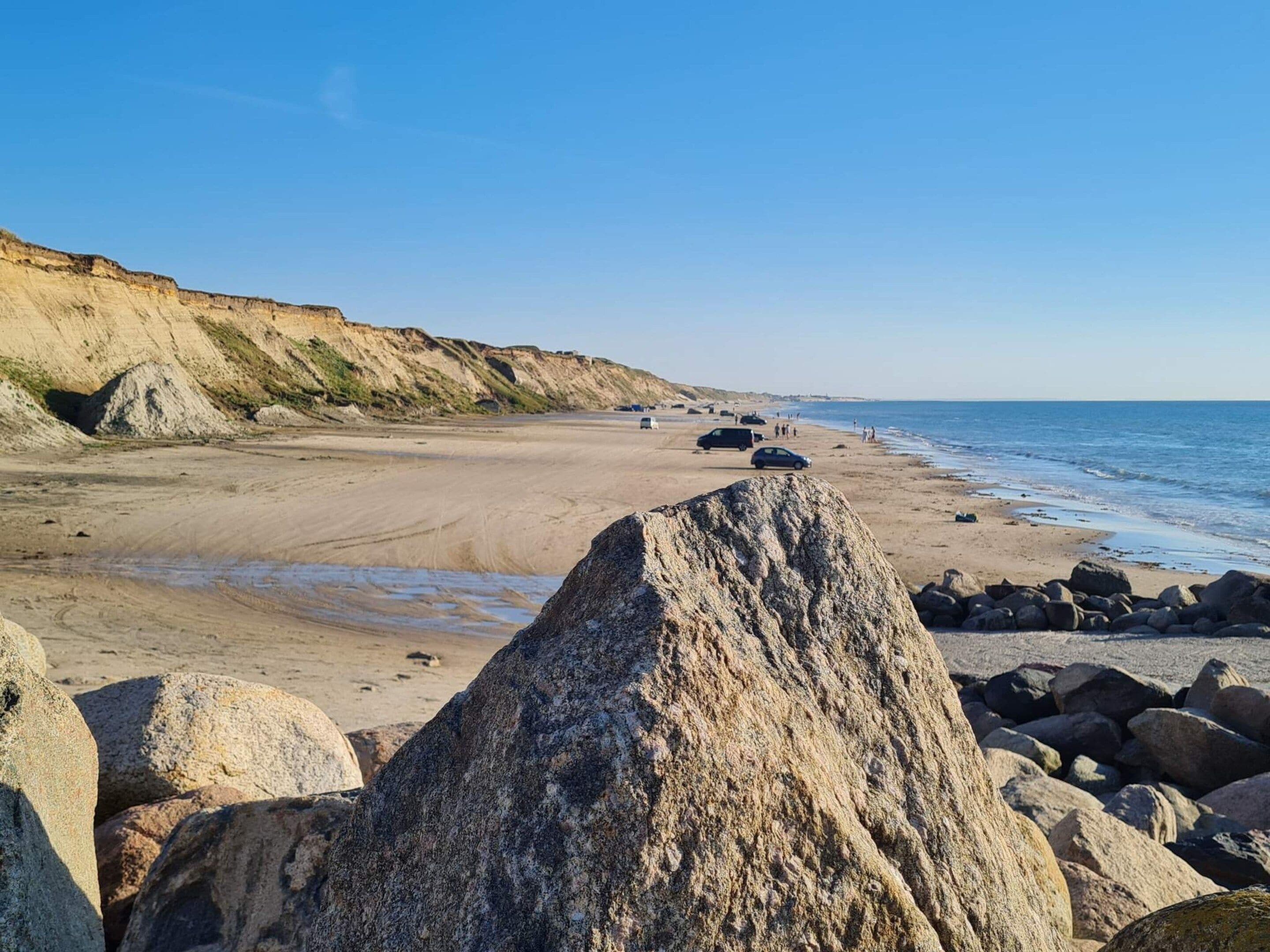 A beach with large rocks in the foreground, a sandy shore, and a cliff in the background.