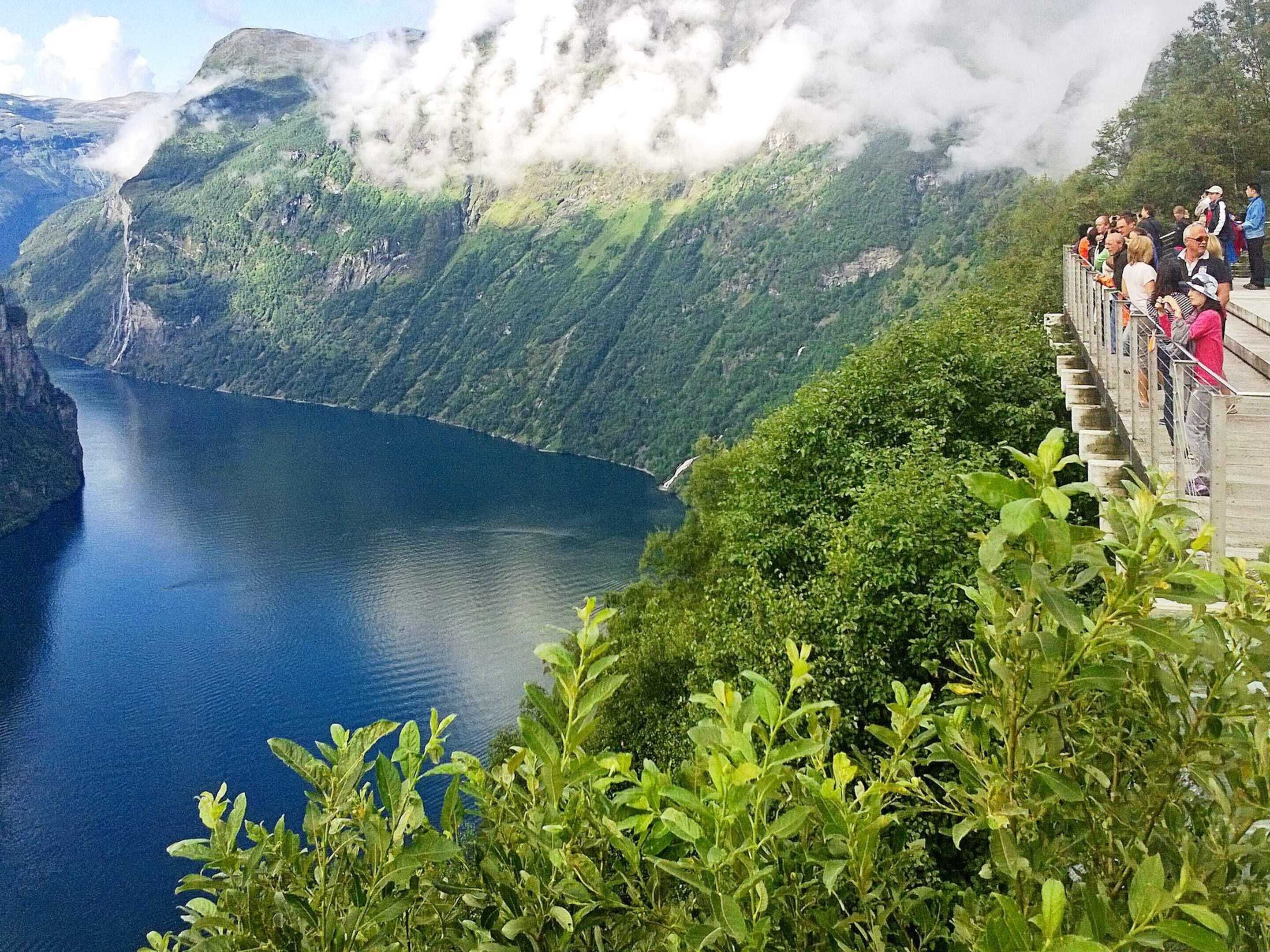 Ein Aussichtspunkt mit Blick auf einen Fjord und Berge, eine hölzerne Aussichtsplattform und eine Menschenmenge.