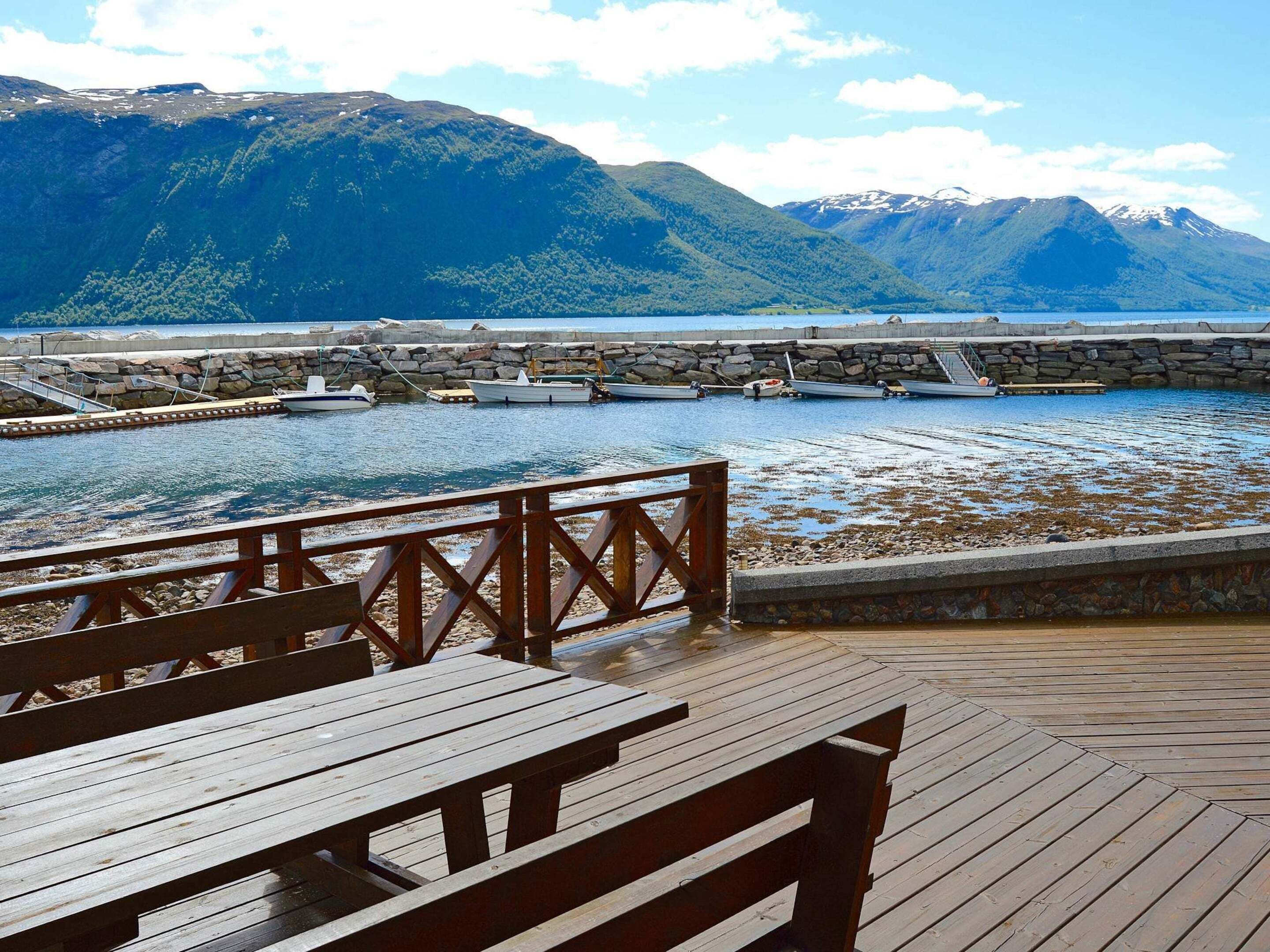 A wooden deck with benches overlooking a calm body of water, a stone pier, and mountains in the background.