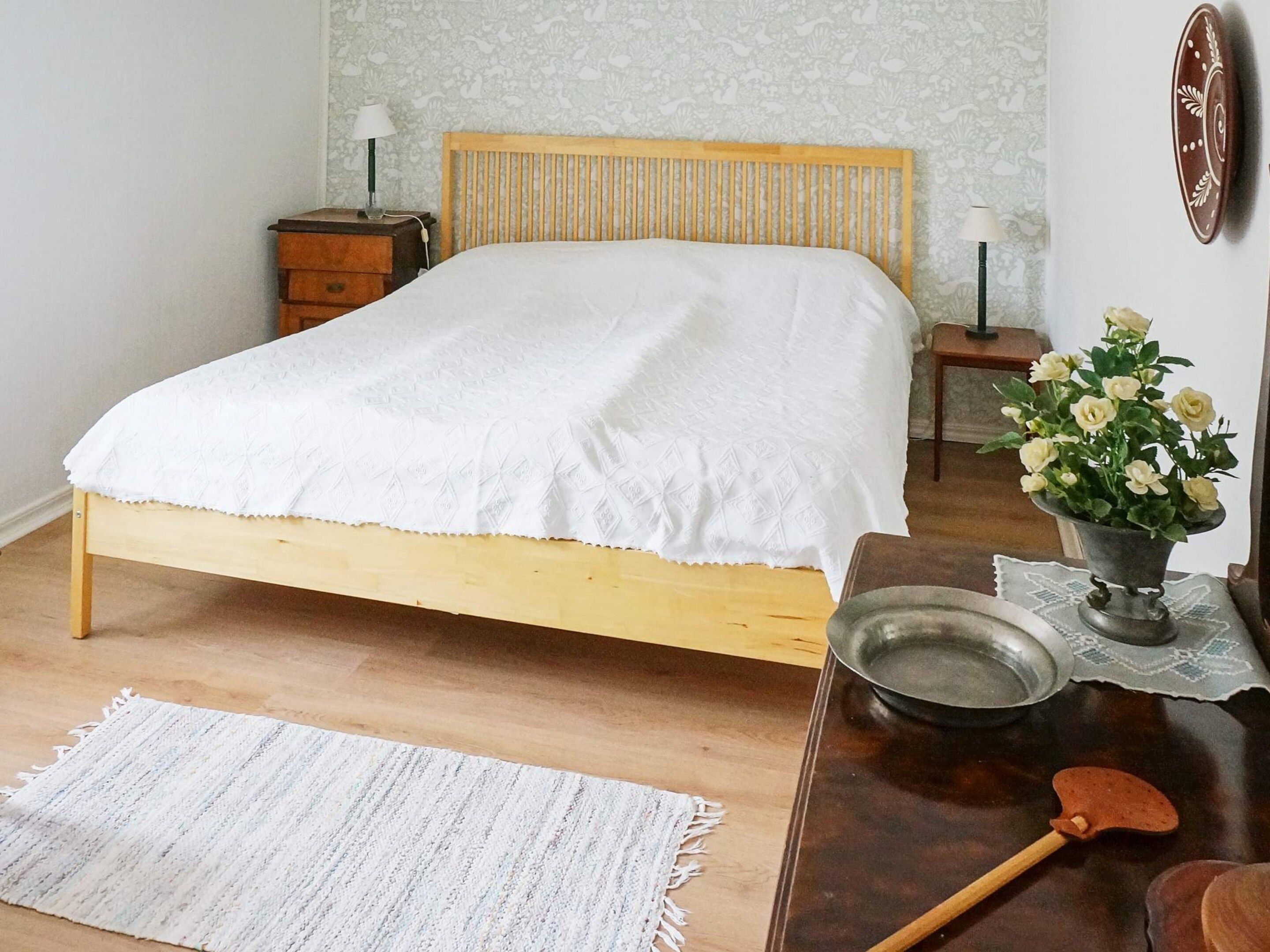 A bedroom with a wooden bed, bedside tables, a nightstand, a bedside lamp, a decorative vase with flowers, and a wooden table with a metal bowl and a wooden spoon.