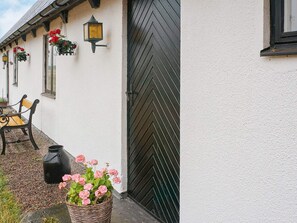 A building with a black chevron-patterned door, a bench, hanging flower baskets, and a wicker basket with pink flowers.