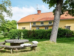 A yellow building with a red-tiled roof, surrounded by greenery and a stone bench with cushions. - Holiday Home in Hova (Hova)