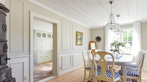 A dining room with a wooden table, white chairs, and a chandelier. There is a kitchen visible through an open doorway. - Holiday Home in Hova (Hova)