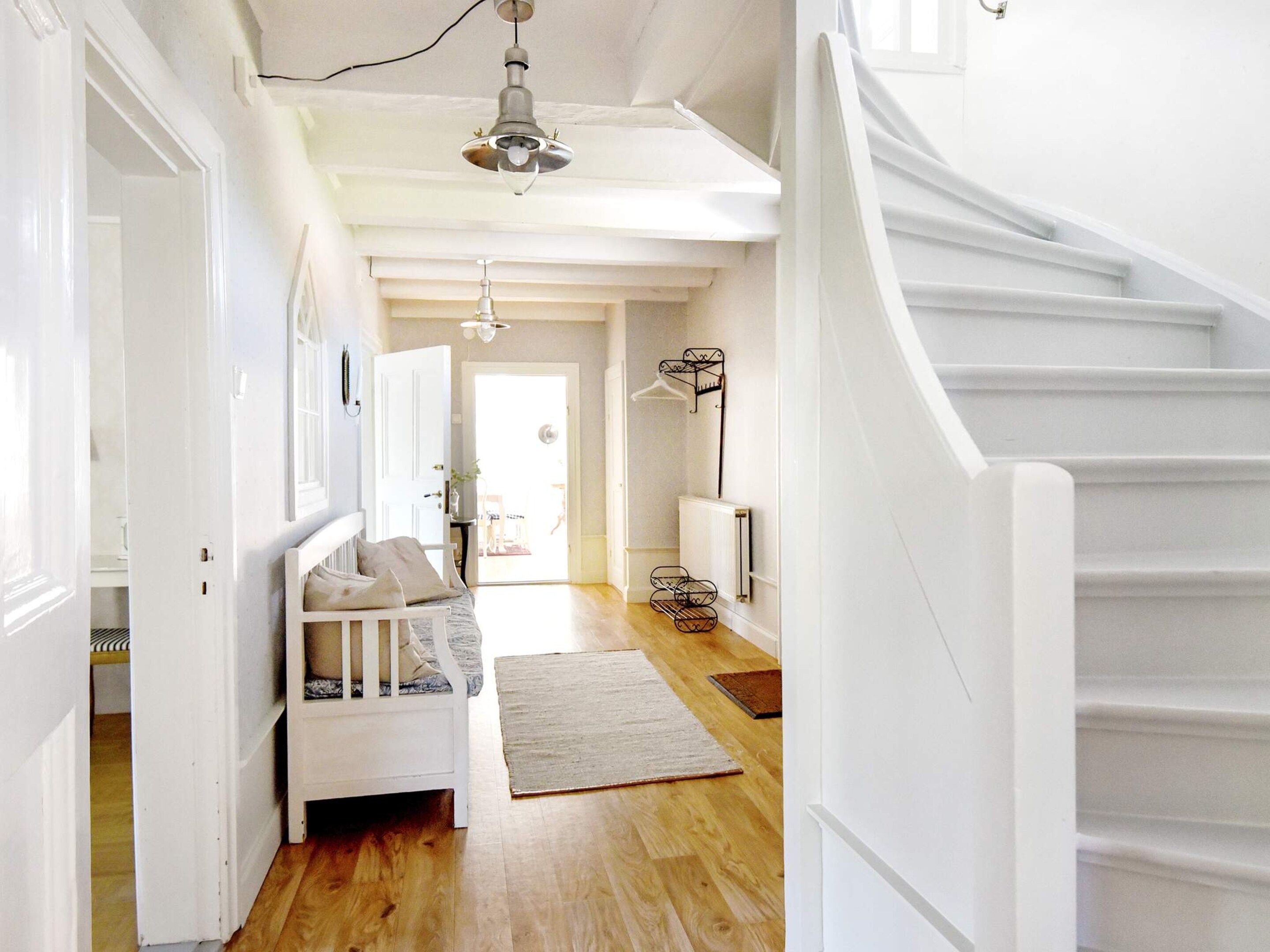 A spacious hallway with a white staircase, a wooden floor, and a bed with a white headboard.