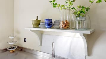 A kitchen with a white countertop, a stainless steel sink, and a shelf above holding a mortar, blue bowls, corks, and a plant.