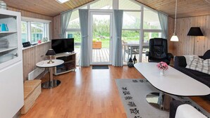 Een gezellige woonkamer met een houten plafond, een televisie, een bank, een eettafel en uitzicht op de omgeving door grote ramen.