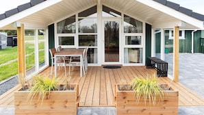 Een overdekt buitengebied met een houten terras, een tafel met stoelen en plantenbakken met groen.
