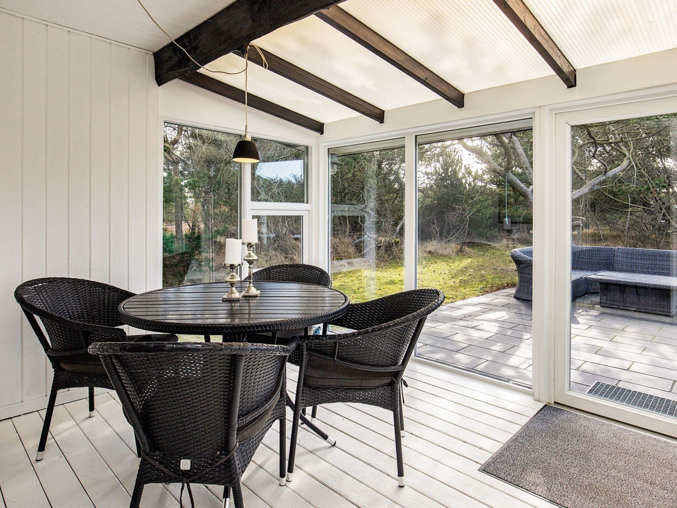 Une terrasse couverte avec une table ronde et des chaises en osier, de grandes portes vitrées et une vue sur l’extérieur.