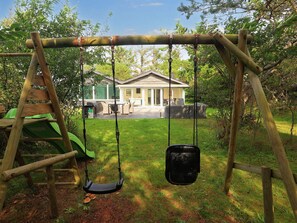 A wooden swing set with a black bucket seat and a green slide, set against a house with a patio and outdoor furniture. - 4 Star Holiday Home in Vejers Strand (Vejers Strand)