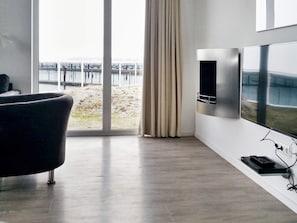 A modern living room with a large TV, a sofa, and a view of a waterfront.