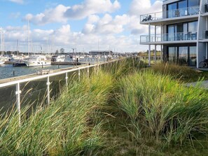 A marina with boats docked, a modern building with balconies, and a grassy area with tall grasses. - Holiday Home in Wendtorf (Wendtorf)