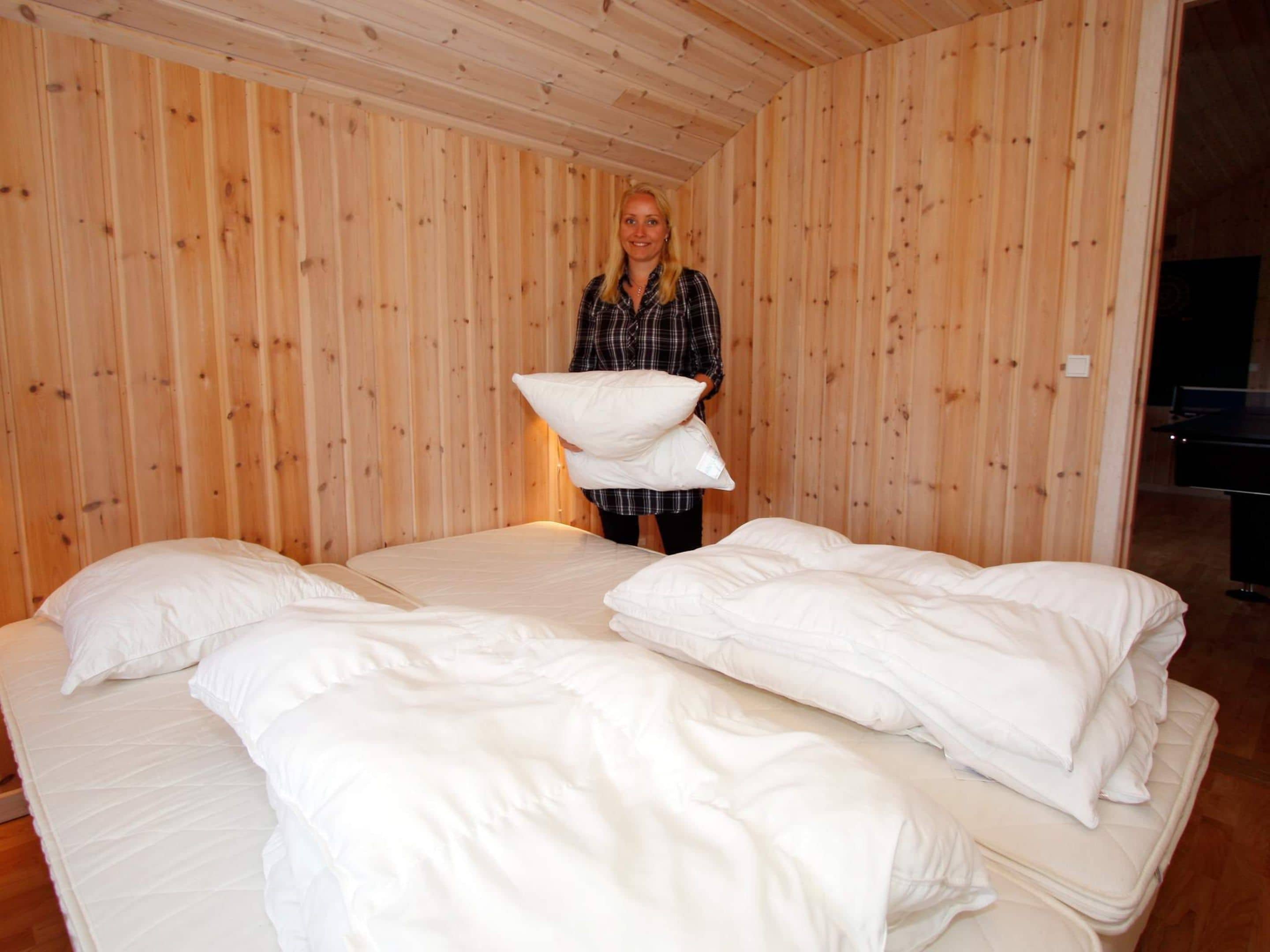 Een kamer met houten paneelwanden, een bed met wit beddengoed, en een persoon die kussens vasthoudt.