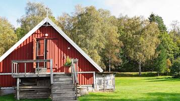 En röd lada med en trätrappa som leder upp till en veranda, omgiven av grönt gräs och träd.