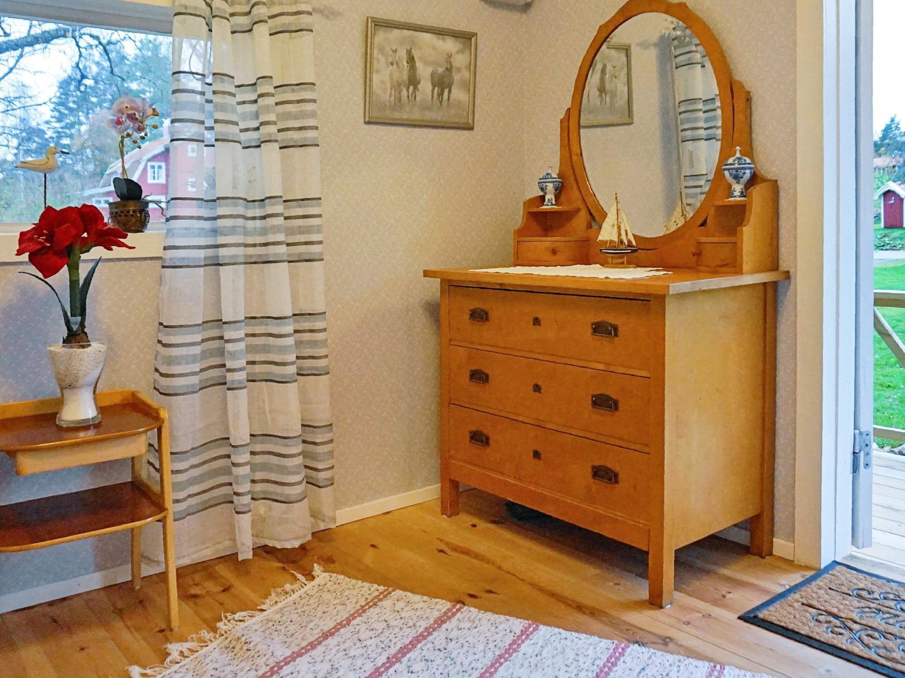 Une pièce comprenant une commode en bois, un miroir, un rideau à rayures et un vase avec des fleurs rouges.