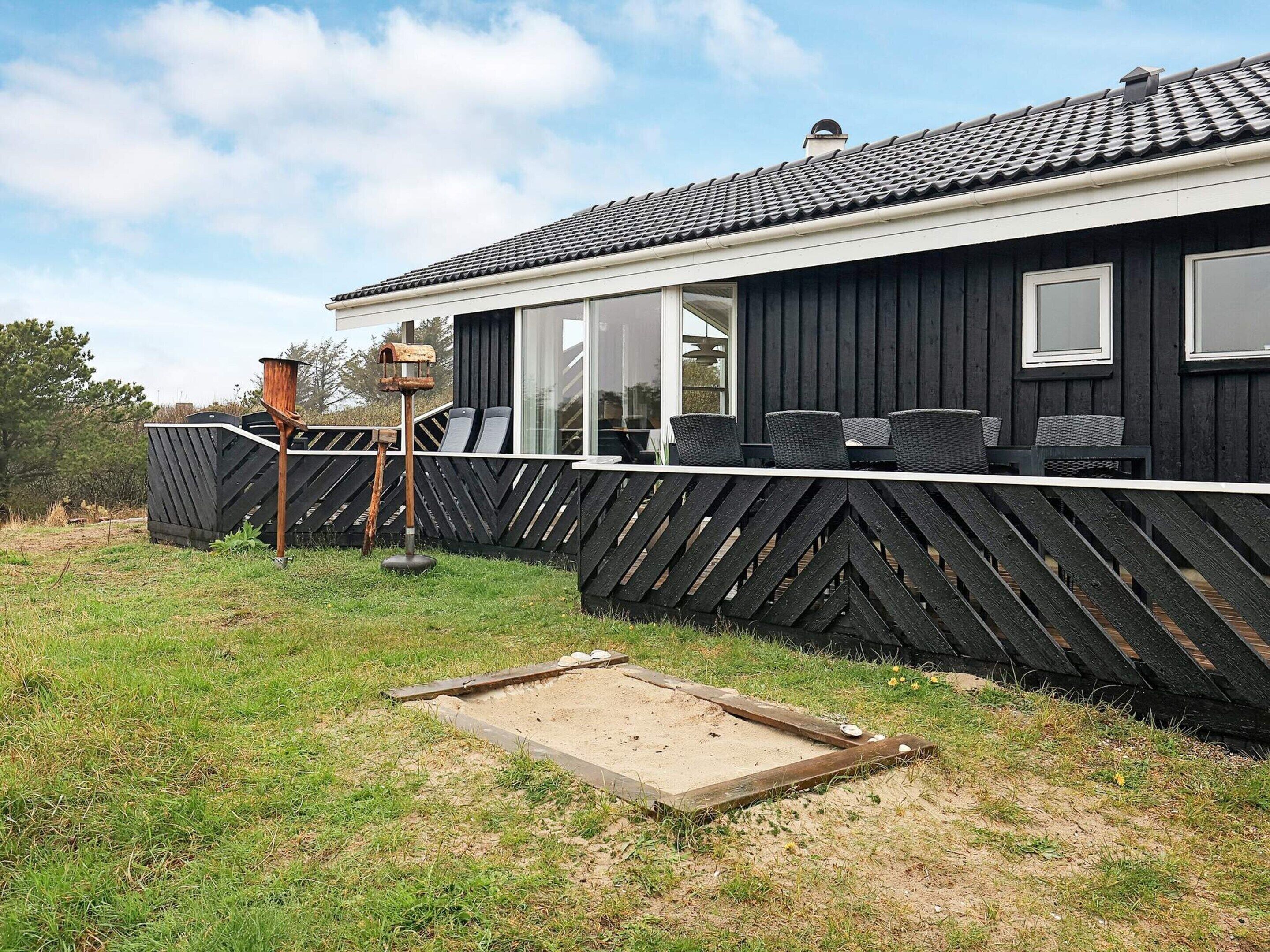 A black wooden building with a tiled roof, a fenced outdoor area with chairs, and a small wooden structure.