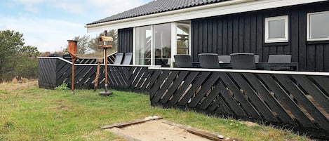 A black wooden building with a tiled roof, a fenced outdoor area with chairs, and a small wooden structure.