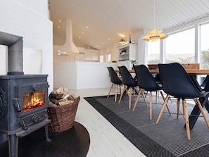 A modern interior with a wood-burning stove, a dining area with chairs, and a view through large windows. - 4 Star Holiday Home in Blokhus (Blokhus)