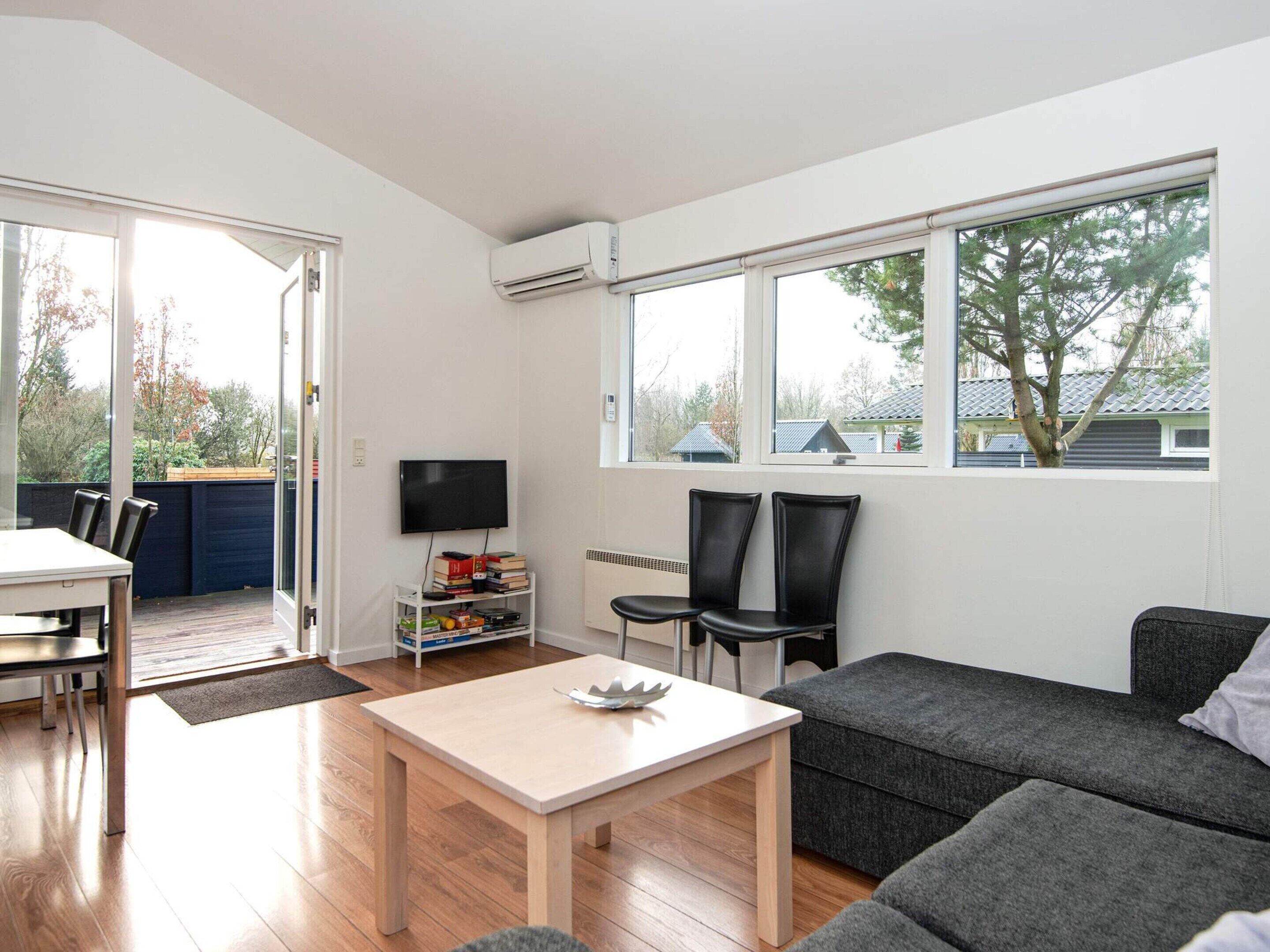 A modern living room with a sofa, a table, and a TV. There is a window with a view of trees and houses.