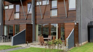 A modern two-story building with large windows, a patio with outdoor furniture, and a small garden area.