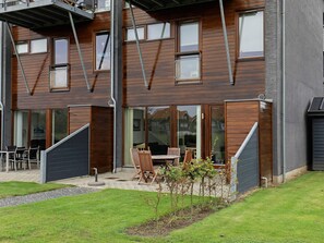 A modern two-story building with large windows, a patio with outdoor furniture, and a small garden area.