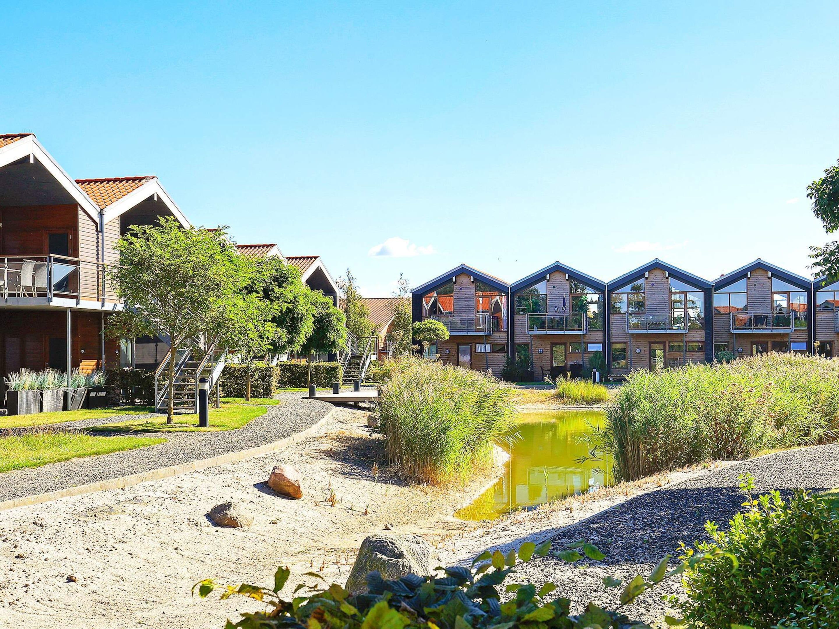 A row of modern wooden cabins with balconies, a gravel path, and a small pond.