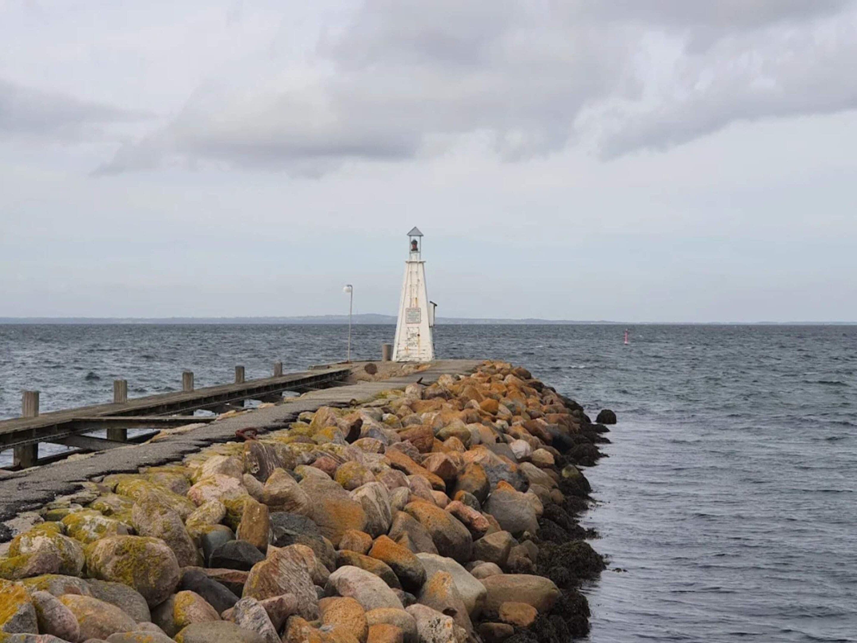 A lighthouse on a rocky pier extending into the sea, with a cloudy sky overhead.
