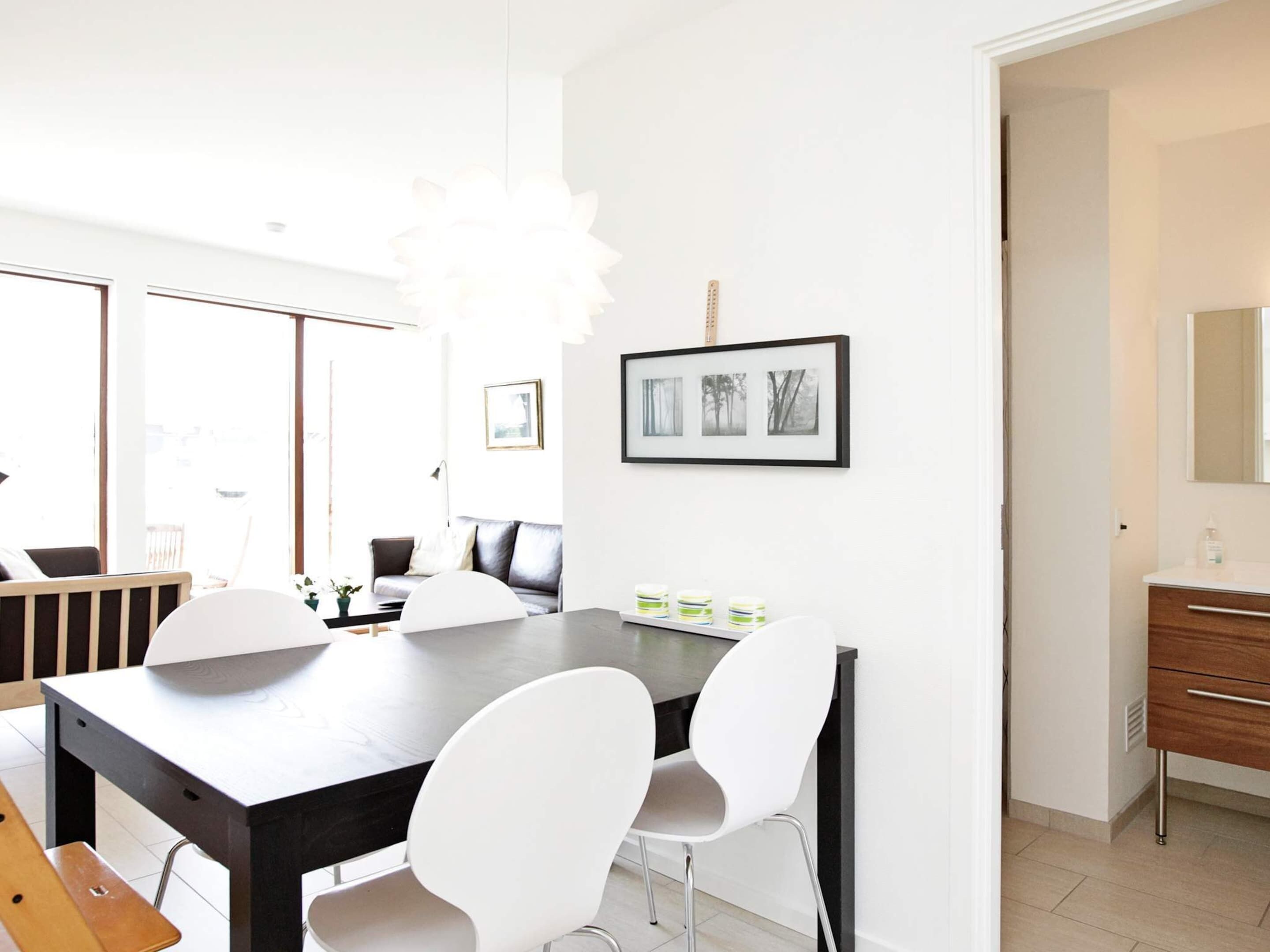 A modern dining area with a dark wooden table, white chairs, and a chandelier. There are framed pictures on the wall and a wooden cabinet with a mirror.