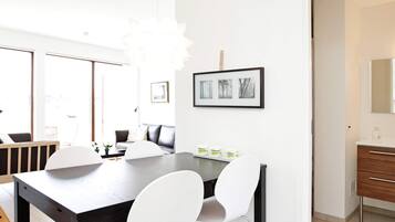 A modern dining area with a dark wooden table, white chairs, and a chandelier. There are framed pictures on the wall and a wooden cabinet with a mirror.