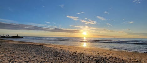 A beach with footprints leading to the water, a clear sky, and the sun setting or rising.