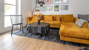 Un salon avec un canapé, une table et une chaise. Les murs sont ornés de tableaux encadrés et il y a une fenêtre d’où l’on peut profiter de la vue.
