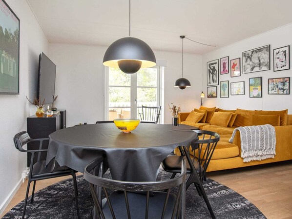 Een moderne woonkamer met een ronde eettafel, zwarte stoelen, een gele bank en zwarte hanglampen.