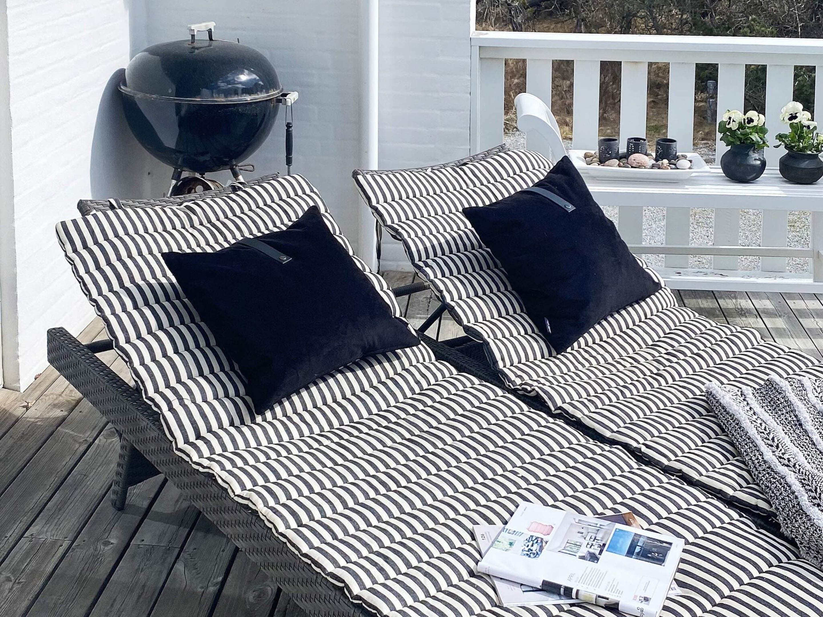 Two striped lounge chairs with black cushions on a wooden deck, a barbecue grill, and a white railing with potted plants.