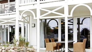 A covered outdoor seating area with wicker chairs and a table, surrounded by a stone wall and glass doors. - Holiday Home in Grossenbrode (Grossenbrode)