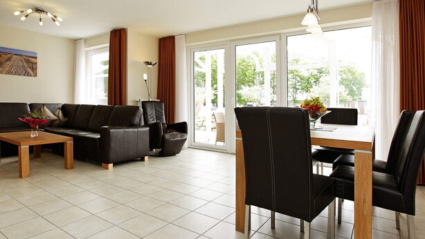 A modern living room with a sofa, a dining table with chairs, and a large window with curtains. - Holiday Home in Grossenbrode (Grossenbrode)